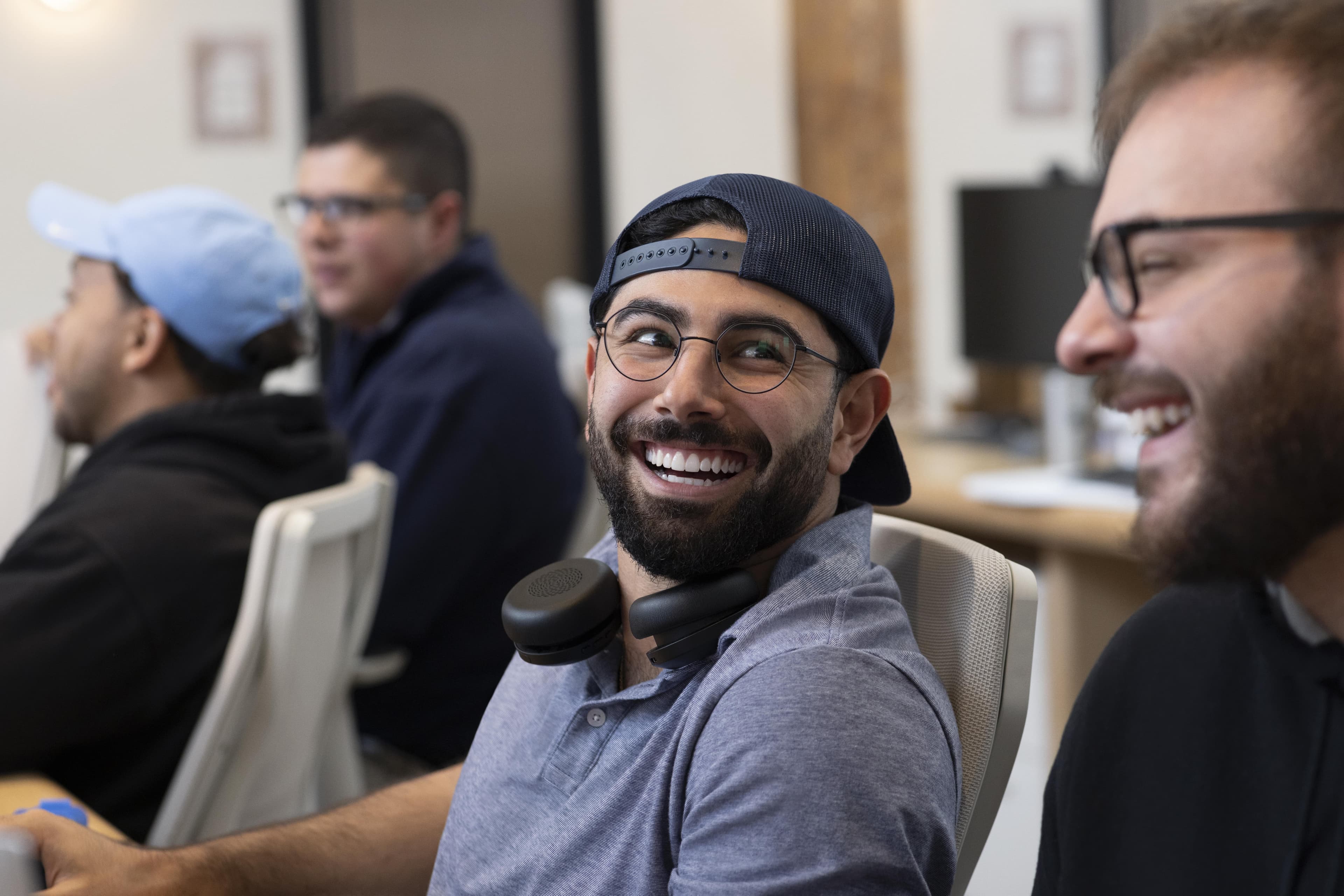 A man with a hat on and head phones around his neck smiling and looking at another smiling man on the edge of the frame