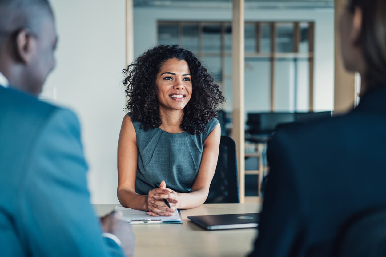 Woman sitting at conference table smiling. She has a clipboard with papers in front of her and is holding a pen. Two people are sitting across from her and are facing away from the camera