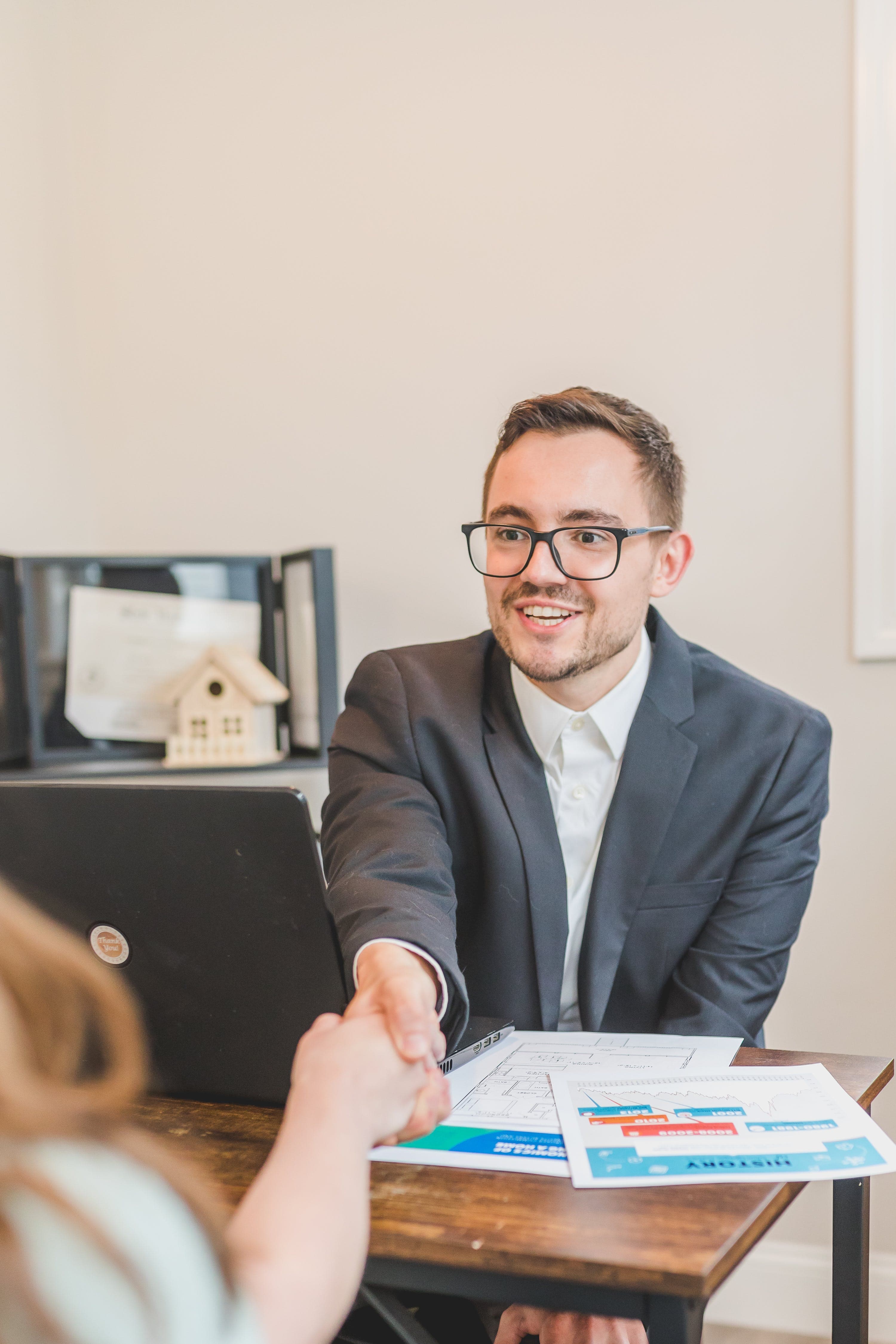 A man sitting at a desk with a laptop and papers on it is shaking hands with someone across the desk who is out of frame